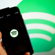 A hand holds a smart phone up, and the screen displays the Spotify logo. In the background, another Spotify logo.